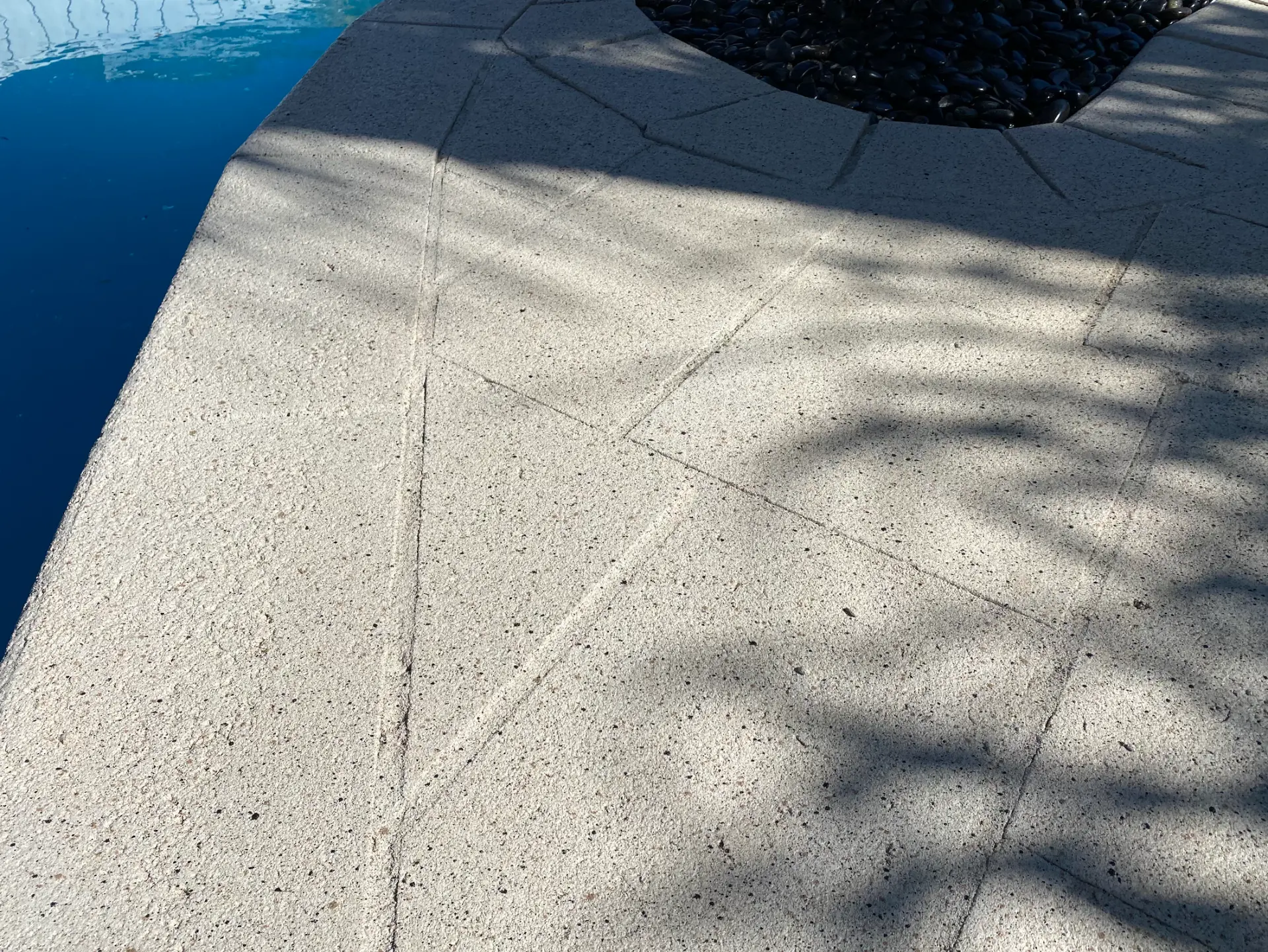 Light grey spray-on concrete pool deck scored into irregular flagstones beside a bed of black river stones at a Perth home